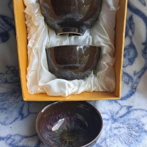 May include: A set of three dark brown ceramic tea bowls with a glossy finish. Two bowls are nestled inside a yellow box lined with white satin fabric, while the third bowl is positioned in front, revealing a dark interior with a floral pattern.