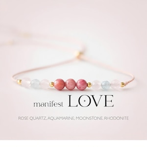 May include: A delicate bracelet featuring rose quartz, aquamarine, moonstone, and rhodonite beads. The bracelet has a light pink cord with gold accents and the text "manifest LOVE". A beautiful piece of jewelry.
