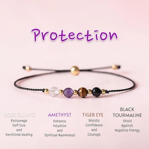 May include: A black cord bracelet with gold beads and various gemstone beads, including rose quartz, amethyst, tiger eye, and black tourmaline. The word "Protection" is written in purple script above the bracelet.
