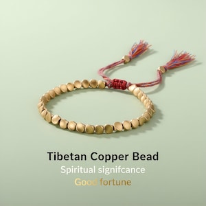 Peut inclure: Un bracelet tibétain en cuivre doré avec un cordon et des pompons rouges et roses. Le bracelet est composé d'une série de perles plates et rondes. Le texte sur l'image indique "Tibetan Copper Bead", "Spiritual significance" et "Good fortune".