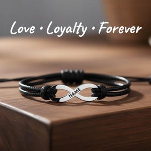 May include: A black cord bracelet featuring a silver-toned infinity symbol charm with the word NAME engraved on it. The text Love • Loyalty • Forever is displayed above the bracelet, which has adjustable sliding knots on the sides.