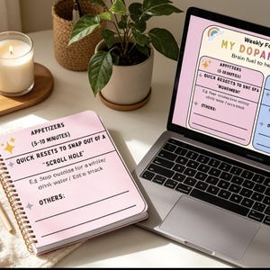 Pode incluir: Um laptop exibe um "Planejador de Foco Semanal" com um design rosa e azul. Um caderno espiral rosa com texto está aberto ao lado do laptop. Uma vela acesa, uma planta e uma caneta também estão na mesa.