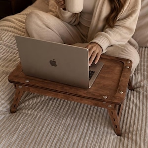 May include: A wooden laptop bed tray with folding legs. The tray is a warm brown color and has metal accents. A silver laptop is on the tray. The scene includes a person sitting on a bed with a mug.