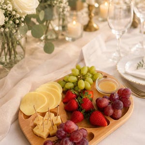 May include: A wooden serving platter with sliced cheese, cubed cheese with toothpicks, green and red grapes, strawberries, and a small bowl of honey. The platter is on a white tablecloth, with flowers and candles in the background.