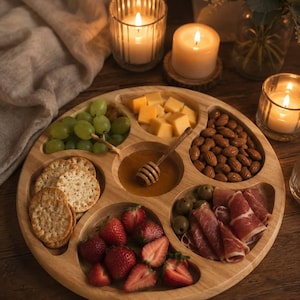 Puede incluir: Una bandeja redonda de madera con varios aperitivos: uvas verdes, cubos de queso, almendras, aceitunas, prosciutto en rodajas, fresas, galletas saladas y miel. Velas encendidas y vegetación en el fondo.