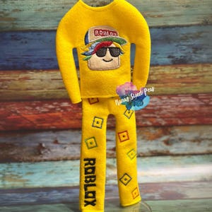 May include: A yellow felt outfit featuring a Roblox-themed design. The top has an embroidered image of a character wearing a hat and sunglasses. The pants have the word "ROBLOX" and square patterns. The outfit is displayed on a clear stand.