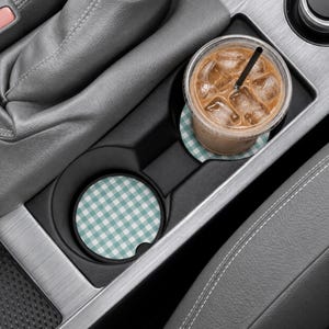 May include: Two circular car cup holder coasters featuring a light teal and white gingham check pattern. One coaster is placed in the empty cup holder, while the other supports a clear plastic cup filled with iced coffee and a black straw.