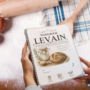 Peut inclure: Un cahier à spirales intitulé "Workbook Levain" avec des illustrations de pain et d'un pot. Le livre est tenu au-dessus d'une surface en bois saupoudrée de farine, à côté d'œufs, d'un rouleau à pâtisserie et d'une cuillère en bois. Le livre porte le texte "Carnet de Fermentation Artisanale."