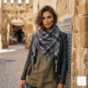 May include: A black and white plaid keffiyeh scarf with fringe trim, worn over an olive green top and a black leather jacket. The scarf features a traditional checkered pattern and is draped around the neck, providing a layered look with the jacket.