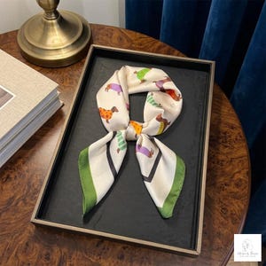 May include: A square silk scarf featuring a cream-colored background with a repeating pattern of small dachshund dogs wearing various colorful sweaters and outfits. The scarf has a distinct dark green border and is tied in a simple knot at the center.
