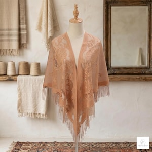Può includere: Uno scialle color pesca con un design floreale in pizzo e finiture con frange. Lo scialle è drappeggiato su un manichino, mostrando il suo motivo intricato e la sua delicata consistenza. Lo sfondo include uno specchio e altri tessuti.