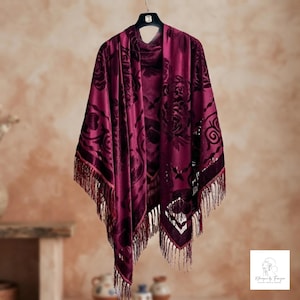 May include: A deep burgundy velvet shawl featuring a tonal floral pattern and a fringe trim along the bottom edge. The garment is displayed on a black hanger and includes a small white logo tag in the bottom right corner with text.