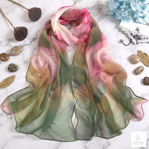 May include: A lightweight, semi-sheer rectangular scarf featuring a gradient pattern of soft pink, muted green, and warm beige tones. The fabric has a delicate, flowing texture with visible folds and a soft, ethereal appearance throughout the piece.