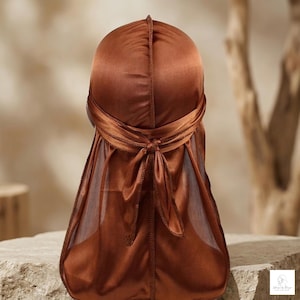Puede incluir: Un durag de satén marrón, diseñado para proteger y peinar el cabello. El durag tiene una cola larga y lazos en la parte posterior, con un detalle de nudo. La tela tiene un brillo sutil, y el diseño es suave y elegante.