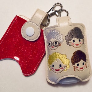 May include: A white hand sanitizer holder with a red glitter pouch attached. The holder features embroidered images of four women with different hairstyles and facial expressions. The holder has a silver key chain attached.