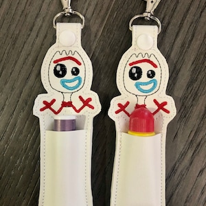 May include: Two white fabric keychains with a cartoon image of the character Forky from the movie Toy Story. Each keychain has a pocket for a lip balm or small tube. One keychain has a purple lip balm in the pocket and the other has a red lip balm.
