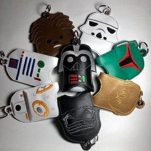 May include: A set of eight Star Wars themed hand sanitizer holders. The holders are made of faux leather and feature embroidered characters from the Star Wars universe. The characters include Chewbacca, Darth Vader, a Stormtrooper, Boba Fett, R2-D2, C-3PO, and two other characters. The holders are all attached to key rings.