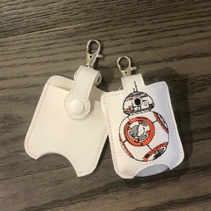 May include: Two white leather hand sanitizer holders with a silver clip. One holder features a black and white embroidered image of a Star Wars character, BB-8.