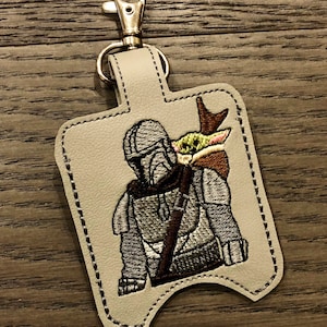 May include: A gray leather keychain with a silver clip. The keychain features an embroidered image of a Mandalorian character from Star Wars holding a small green alien creature.