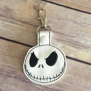 May include: White keychain featuring a stitched Jack Skellington face with black eyes and a stitched smile. The keychain has a silver clasp and a white snap closure.