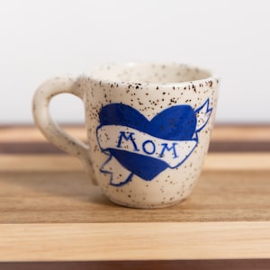 May include: A cream-colored ceramic mug with a speckled finish featuring a blue heart graphic wrapped in a white banner with the word MOM written in blue ink. The mug has a single handle on the left side and a rounded body shape.