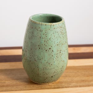 May include: A speckled, light green ceramic vase with a rounded base and a slightly flared top. The vase has a smooth, glossy finish and is approximately 5 inches tall. The surface is covered in small, dark brown speckles.