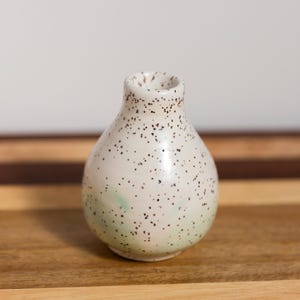 May include: A small, speckled ceramic vase with a rounded body and a narrow neck. The vase is primarily off-white with small, dark brown speckles and a hint of pale green near the base. It sits on a wooden surface.