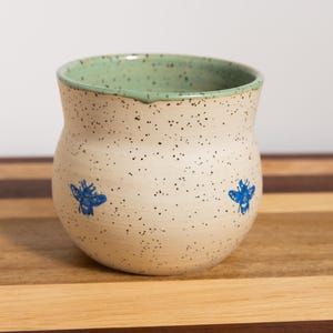 May include: A speckled ceramic mug with a light green interior and two blue bee illustrations. The mug has a rounded shape and sits on a wooden surface.