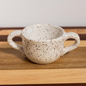 May include: A small, round ceramic cup featuring a speckled off-white glaze with brown flecks throughout. The cup has two curved handles on opposite sides, a wide circular opening, and a smooth, rounded base, creating a symmetrical design.