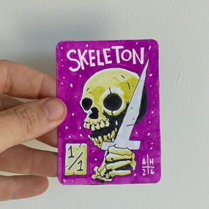May include: A rectangular trading card with a vibrant purple background featuring a yellow skull holding a white dagger. The text SKELETON is printed at the top, with a 1/1 box in the bottom left corner and the initials AH 2/6 in the bottom right.