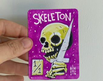 May include: A rectangular trading card with a vibrant purple background featuring a yellow skull holding a white dagger. The text SKELETON is printed at the top, with a 1/1 box in the bottom left corner and the initials AH 2/6 in the bottom right.