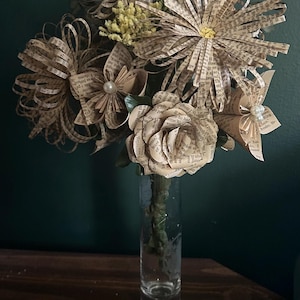 May include: A bouquet of handcrafted flowers made from vintage book pages, featuring various petal shapes and sizes. Some flowers are accented with small white faux pearls at their centers. The arrangement is displayed in a clear glass vase.