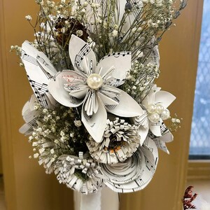 May include: A bouquet of handcrafted paper flowers made from vintage sheet music, featuring white petals with black musical notation. The arrangement includes dried lavender, baby's breath, and central pearl accents, all displayed in a white vase.