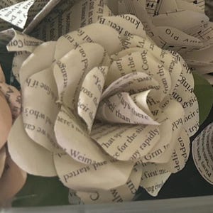 May include: A handcrafted paper flower shaped like a rose, constructed from off-white book pages featuring black printed text. The petals are layered and curled to create a dimensional bloom, with visible snippets of sentences and words throughout.