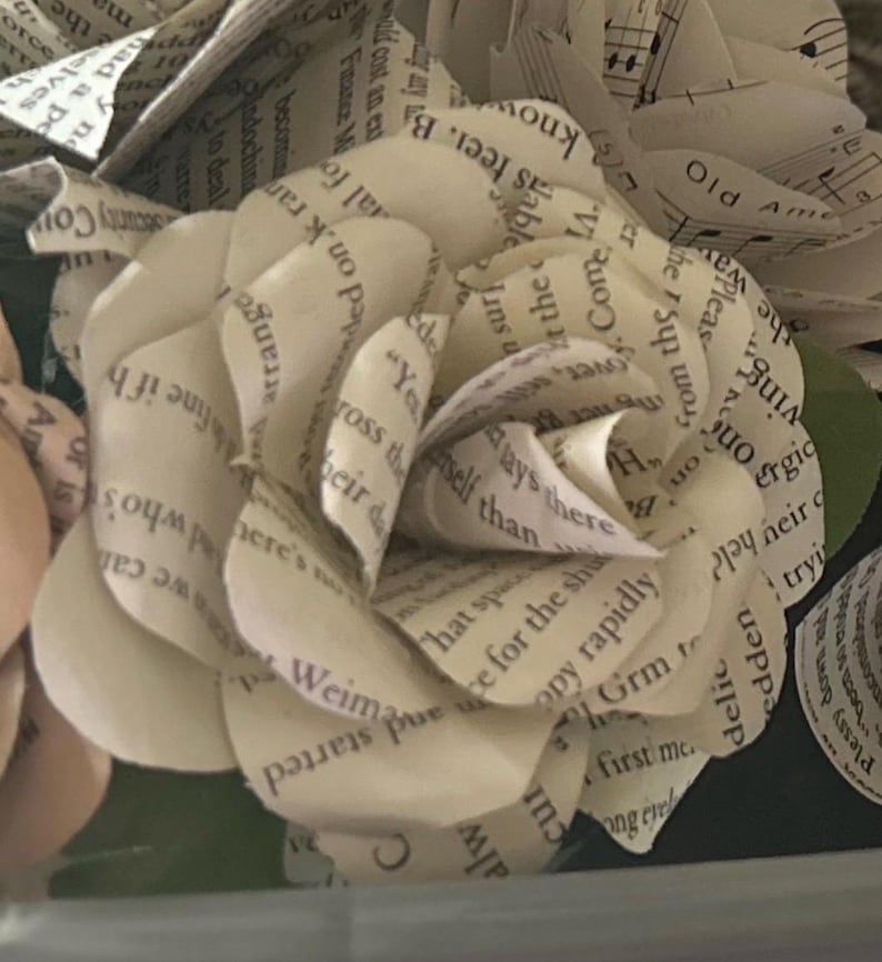 May include: A handcrafted paper flower shaped like a rose, constructed from off-white book pages featuring black printed text. The petals are layered and curled to create a dimensional bloom, with visible snippets of sentences and words throughout.