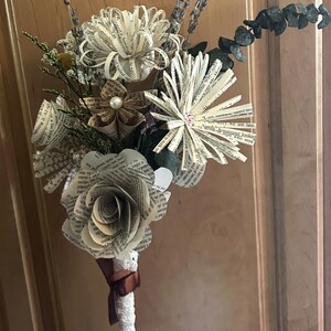 May include: A bouquet of handcrafted flowers made from printed book pages, featuring various bloom styles with text visible on the petals. The arrangement includes accents of dried greenery and small pearls, with a white lace-wrapped stem tied with a brown ribbon.