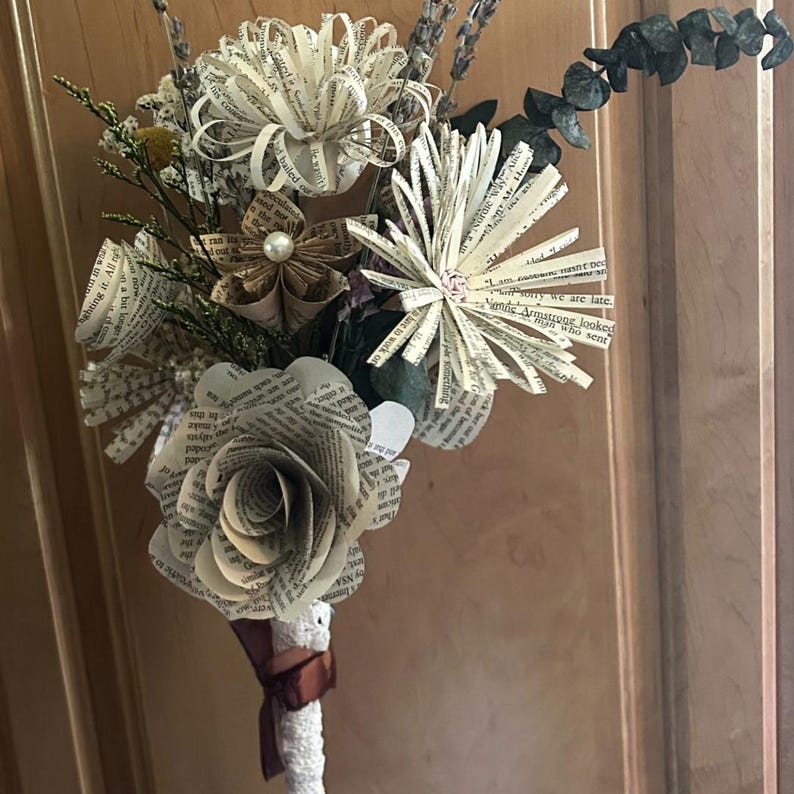 May include: A bouquet of handcrafted flowers made from printed book pages, featuring various bloom styles with text visible on the petals. The arrangement includes accents of dried greenery and small pearls, with a white lace-wrapped stem tied with a brown ribbon.