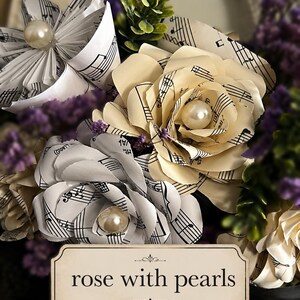 May include: A bouquet of handcrafted paper flowers made from vintage sheet music, featuring cream and white folded petals accented with central faux pearls. The arrangement includes sprigs of purple dried flowers and green foliage.