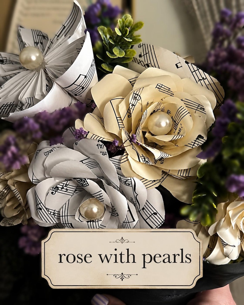 May include: A bouquet of handcrafted paper flowers made from vintage sheet music, featuring cream and white folded petals accented with central faux pearls. The arrangement includes sprigs of purple dried flowers and green foliage.