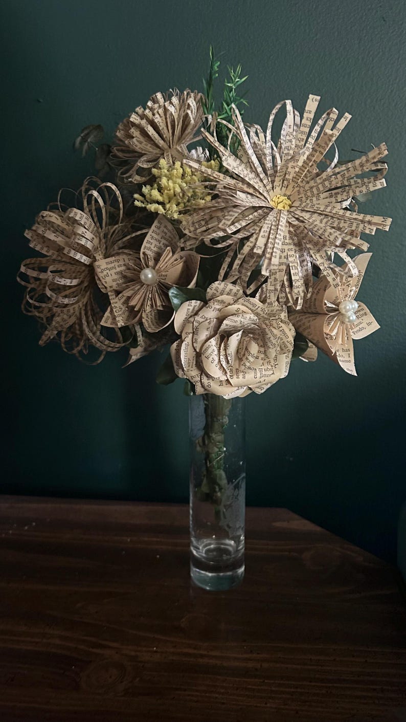 May include: A bouquet of handcrafted flowers made from vintage book pages, featuring various petal shapes and sizes. Some flowers are accented with small white faux pearls at their centers. The arrangement is displayed in a clear glass vase.