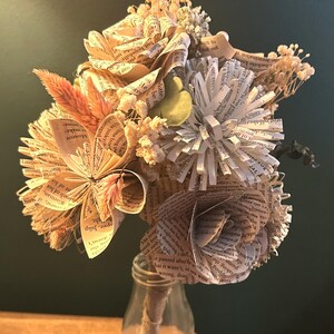May include: A bouquet of handcrafted flowers made from vintage book pages, featuring folded paper roses and star-shaped blooms. The arrangement includes dried beige grasses and small white filler flowers, all secured in a clear glass bottle.