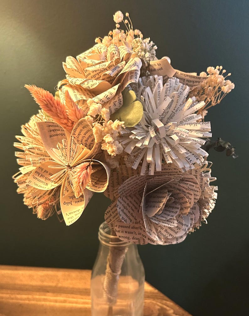 May include: A bouquet of handcrafted flowers made from vintage book pages, featuring folded paper roses and star-shaped blooms. The arrangement includes dried beige grasses and small white filler flowers, all secured in a clear glass bottle.