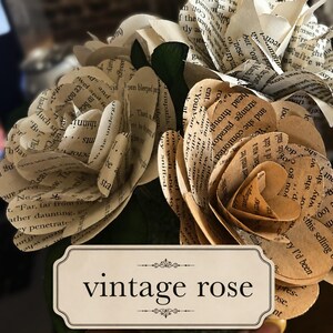 May include: A bouquet of paper flowers crafted from vintage book pages with visible printed text. The flowers feature layered petals in shades of cream and tan. A rectangular label with a decorative border displays the text vintage rose.