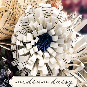 May include: A handcrafted paper flower sculpture featuring layered white strips with black musical staff lines and a dark blue center. The petals are constructed from cut paper, and the base includes a label with the text medium daisy.