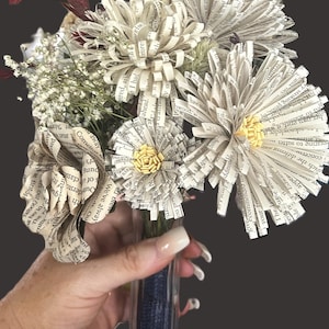 May include: A bouquet of handcrafted flowers made from printed book pages, featuring off-white paper petals with visible black text. The arrangement includes small yellow paper centers, sprigs of white baby's breath, and dark red foliage.