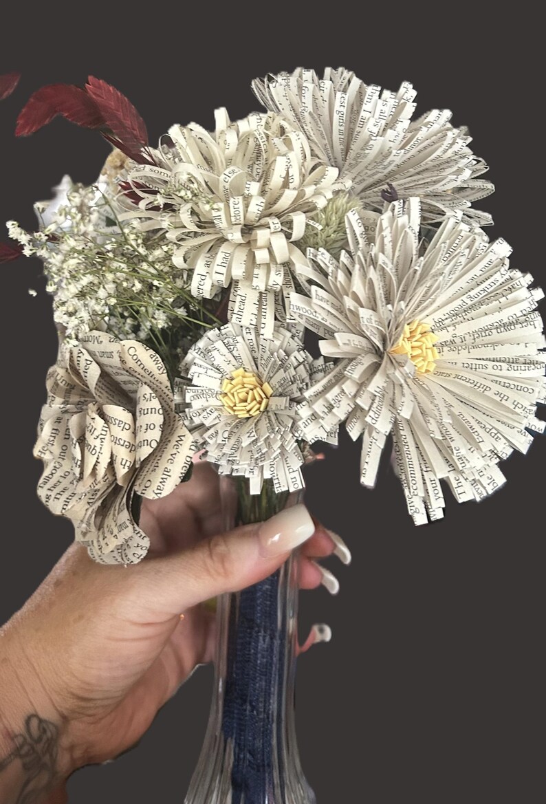 May include: A bouquet of handcrafted flowers made from printed book pages, featuring off-white paper petals with visible black text. The arrangement includes small yellow paper centers, sprigs of white baby's breath, and dark red foliage.