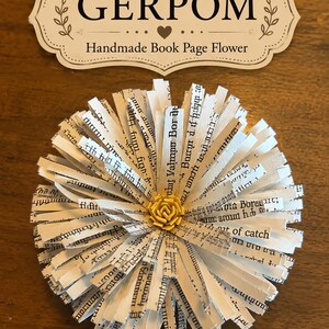 May include: A decorative flower crafted from strips of printed book pages with black text, featuring a small yellow center. Above the flower is a cream-colored label with a decorative border containing the text GERPOM Handmade Book Page Flower.