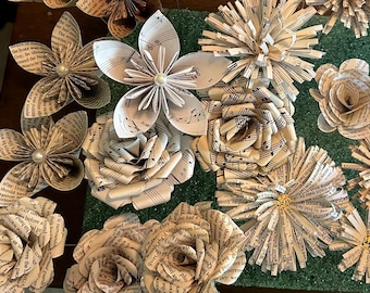 May include: A collection of handcrafted paper flowers made from vintage book pages and sheet music. The flowers feature various petal shapes, some with central white pearl or gold accents, displaying printed text and musical notation throughout.