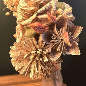 May include: A bouquet of handcrafted flowers made from vintage book pages, featuring folded paper petals with visible printed text. The arrangement includes dried cream-colored baby's breath and small pink feathery stems, all held in a clear glass bottle.