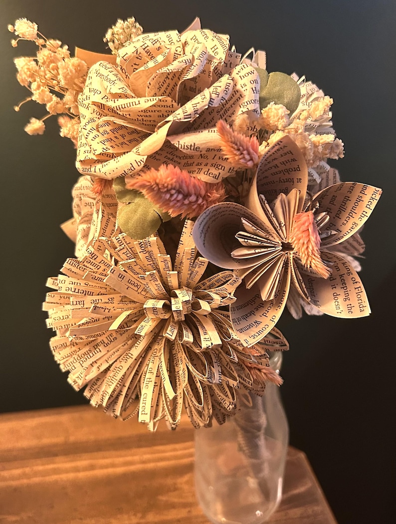 May include: A bouquet of handcrafted flowers made from vintage book pages, featuring folded paper petals with visible printed text. The arrangement includes dried cream-colored baby's breath and small pink feathery stems, all held in a clear glass bottle.